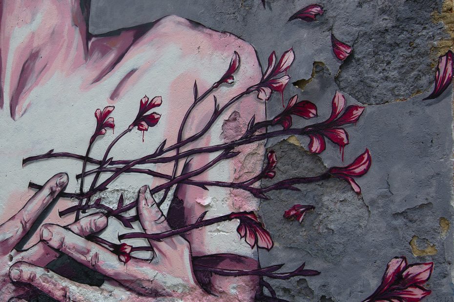 Foto einer Wandmalerei. Zu sehen ist eine kaputte Wand mit einem halben Oberkörper und Händen die rote Blumen halten, die fast wegfliegen.